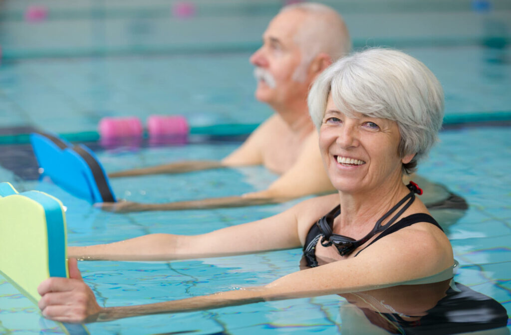 An older adult smiles at the camera while holding a floating board in the pool beside a friend during aquatic therapy